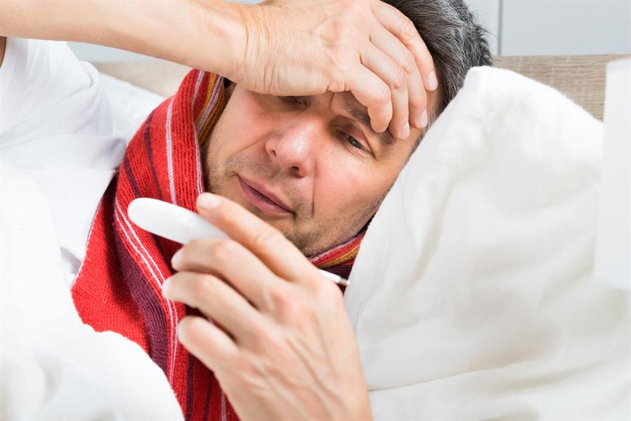 Middle-aged man in bed holding a thermometer in one hand and feeling his forehead with another hand.