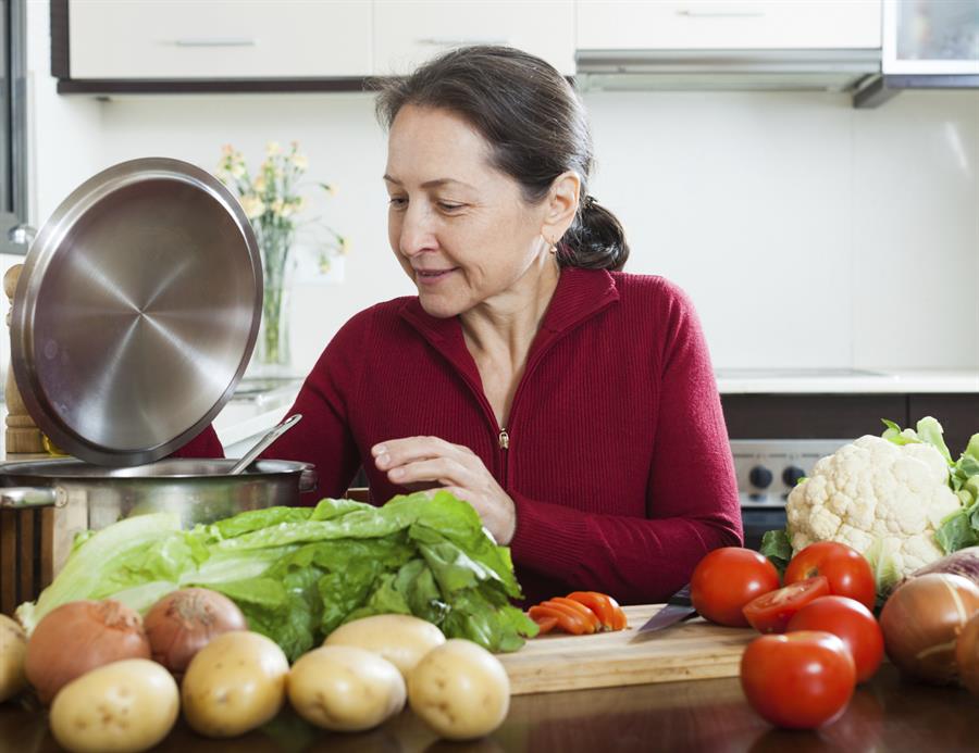 Older woman in kitchen with cooking pot and vegetables.