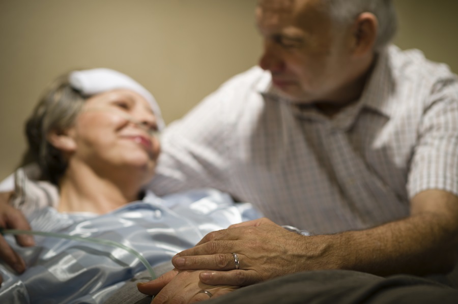 Ailing woman in bed with bandage on her head, man looking at and embracing her, both smiling.