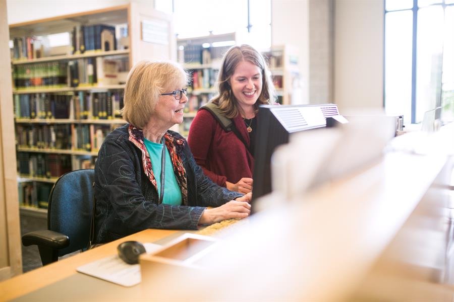 Young librarian teaching a senior woman how to use library computers.