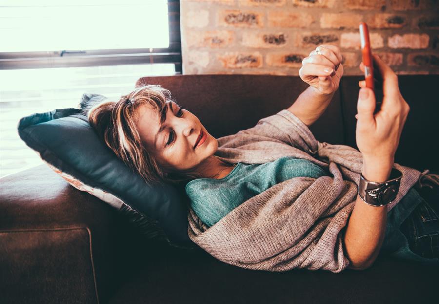 Older woman lying down on a couch and scrolling through a mobile phone.