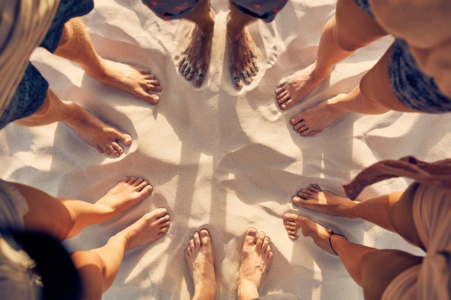 A group of people standing barefoot in the sand in a circle formation with only their legs and/or feet showing.  