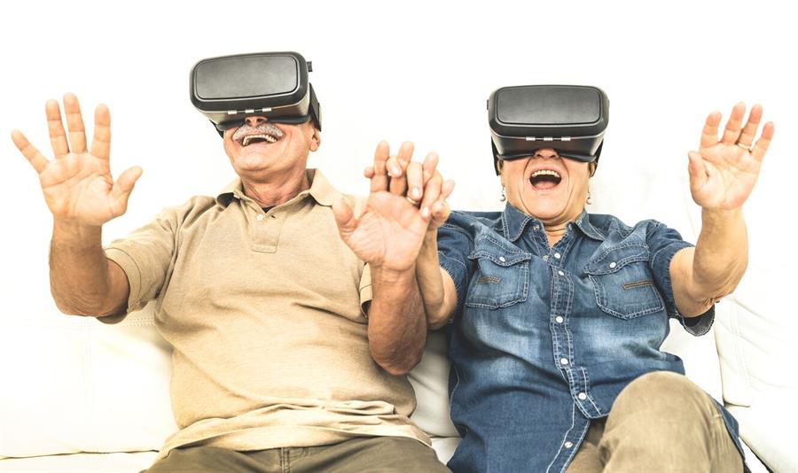 Older couple sitting on a couch with VR headsets on with their hands out in amazement.