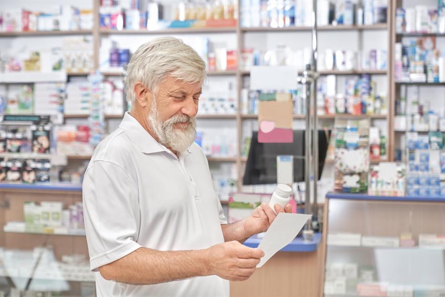 Older adult male standing in a pharmacy while holding a bottle of medication and reading a piece of paper.   