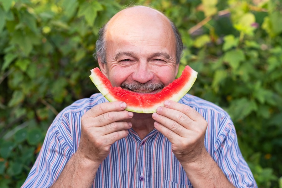 An older adult male standing outside and holding up an eaten slice of watermelon to mimic a smile.
