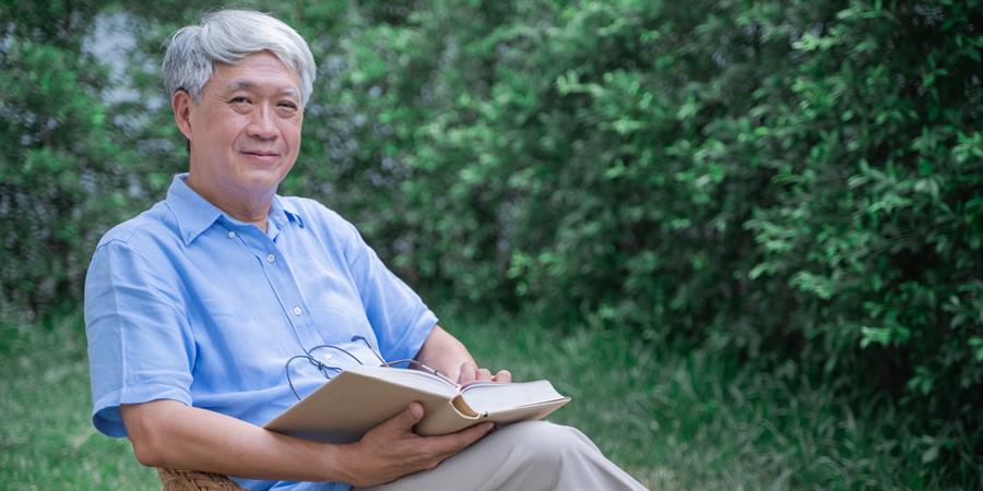 Older adult man sitting on a chair outdoors and reading a book.