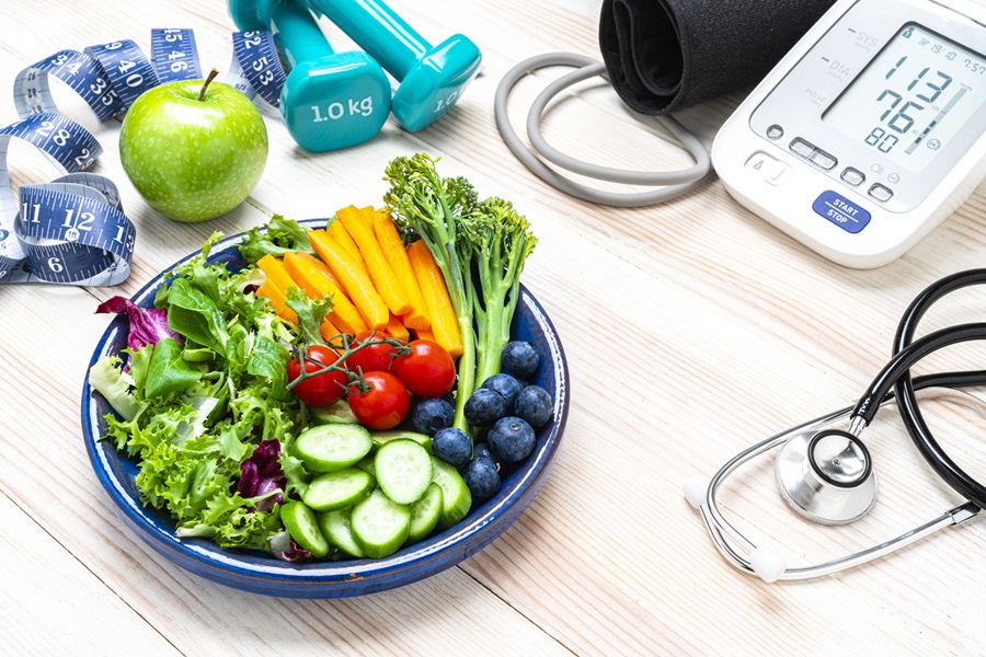 Electronic blood pressure monitor and bowl of fruits, vegetables and dumbbells representing healthy lifestyle choices