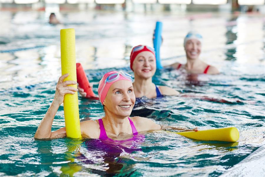 Smiling female in swimwear holding a pool noodle with two more swimming women in an indoor pool.