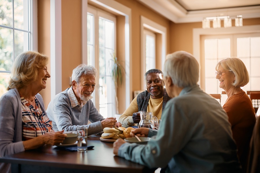 Group of five older adults sitting around a dining room table eating and chatting.