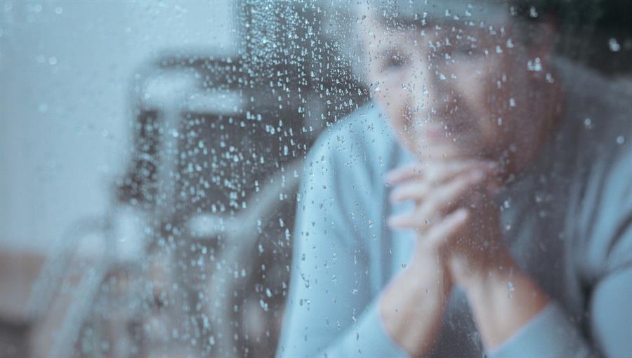 Senior woman sitting with her hands under her chin, while staring outside of a window on a rainy day. A wheelchair is visible in the background. 