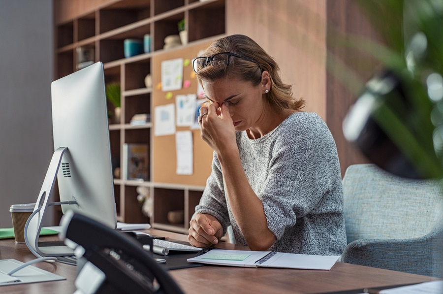 Business woman sitting in front of a computer looking down and pinching the bridge of her nose in stress.