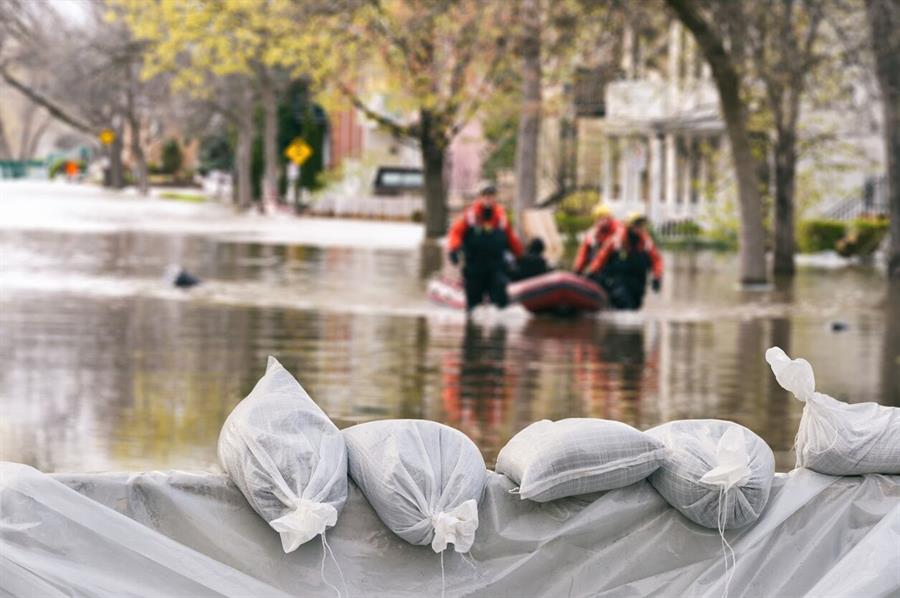 Flood protection sandbags with flooded homes and a rescue team in the background.